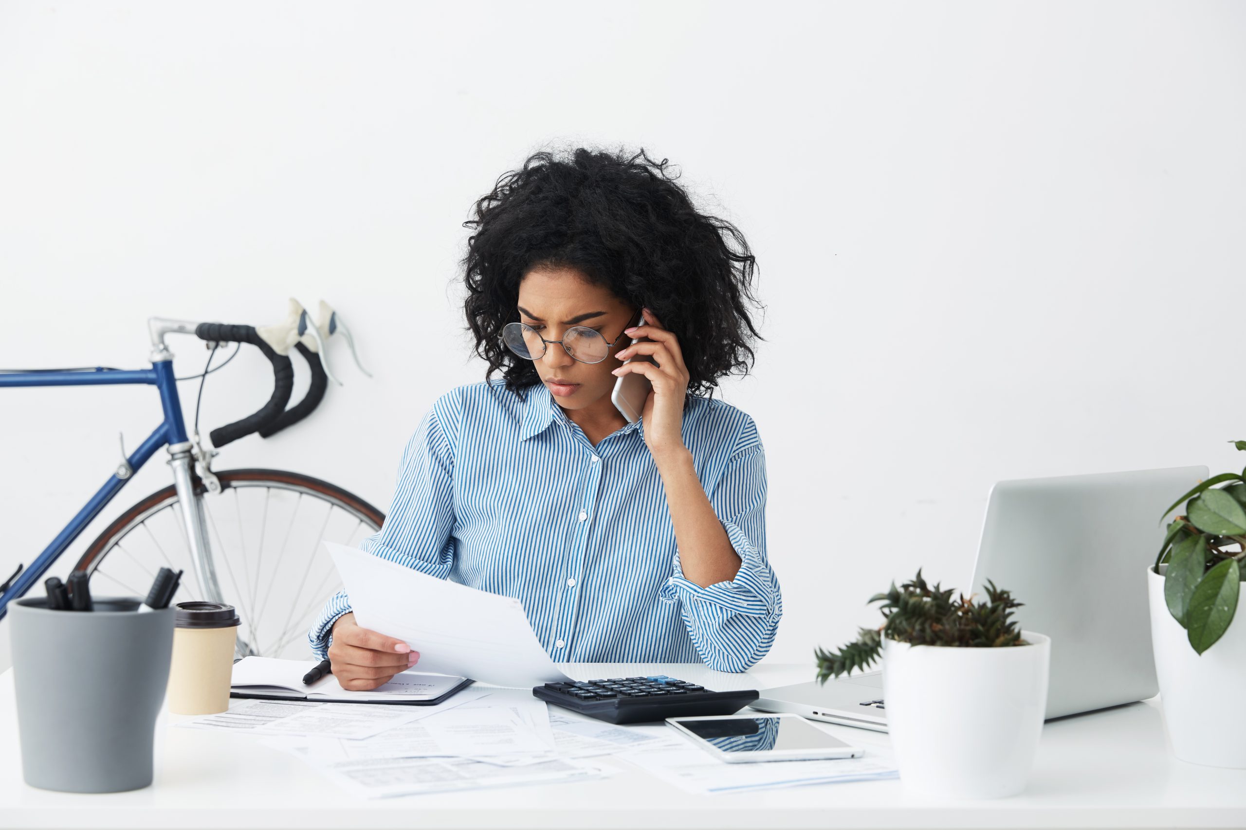 Woman in glasses talks on the phone while reviewing a document at her desk.
