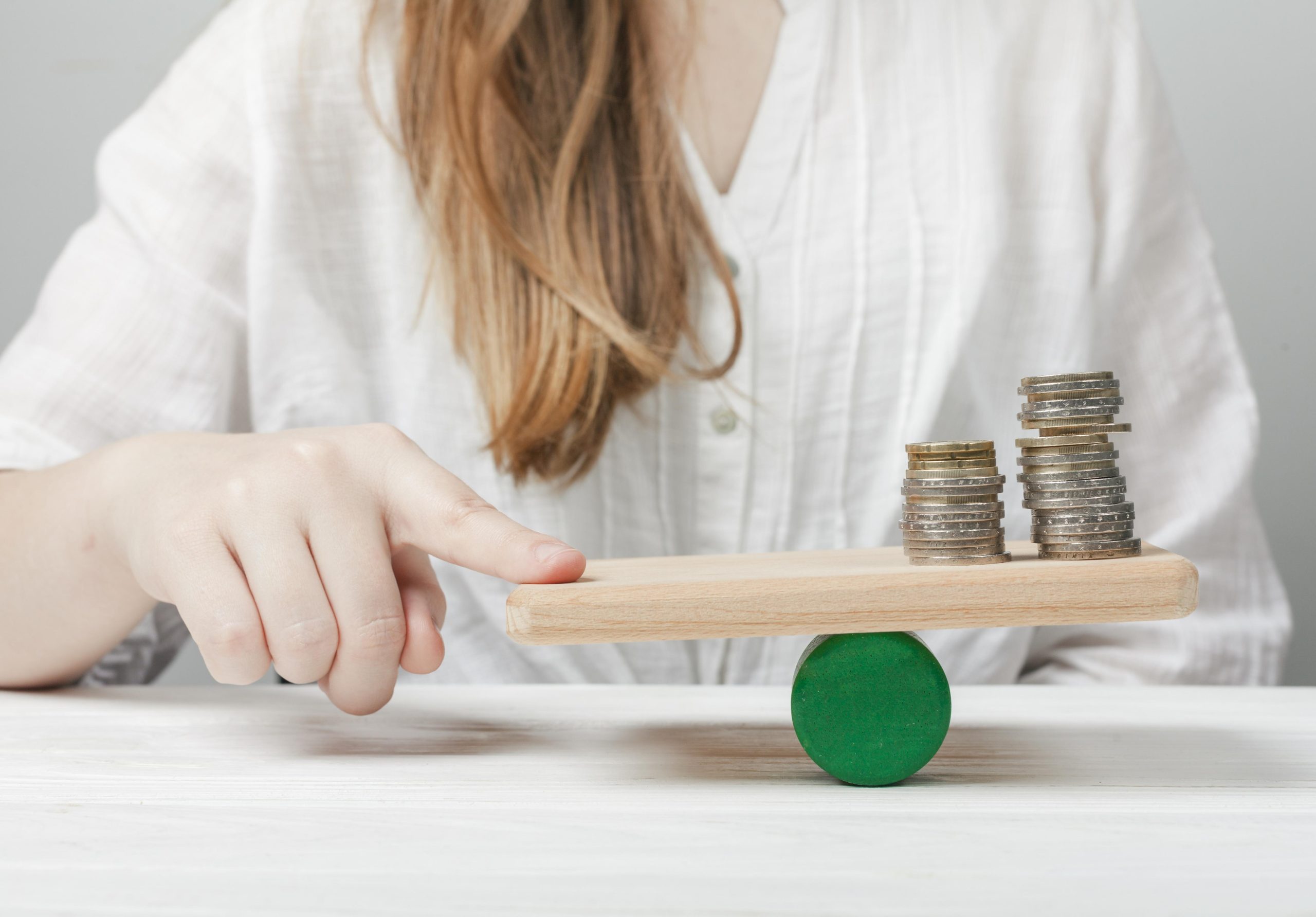 A person's finger gently presses down on a wooden seesaw balancing platform, which holds two small stacks of coins