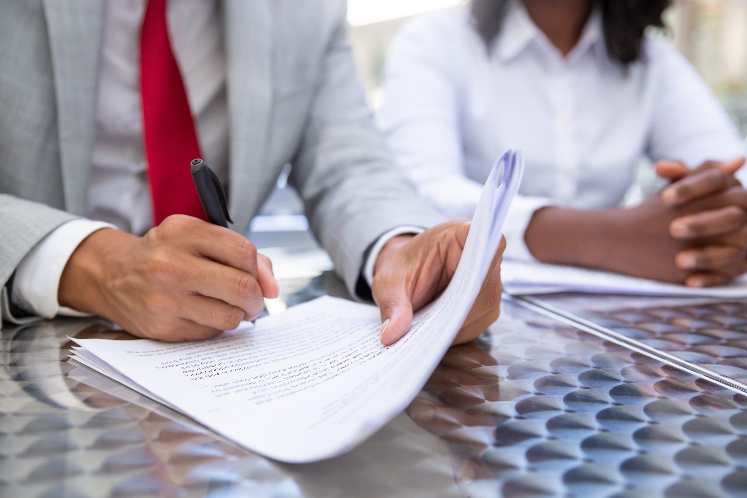 Close-up of a man in a gray suit and red tie signing a document with a black pen, while an unseen person sits across the table.