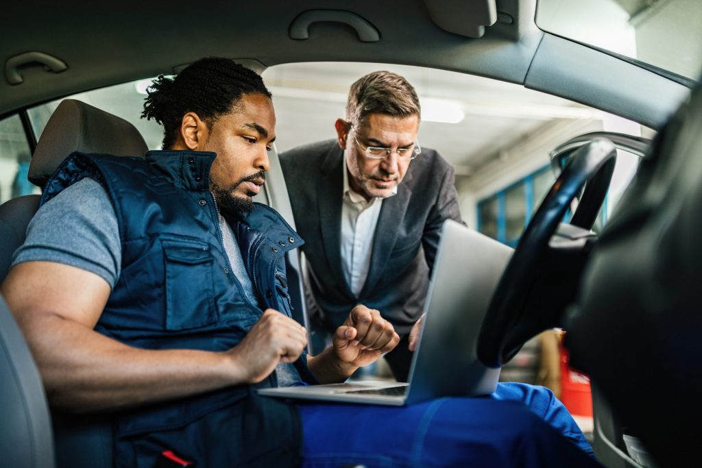 Young black car mechanic using computer with his manager in auto repair shop.
