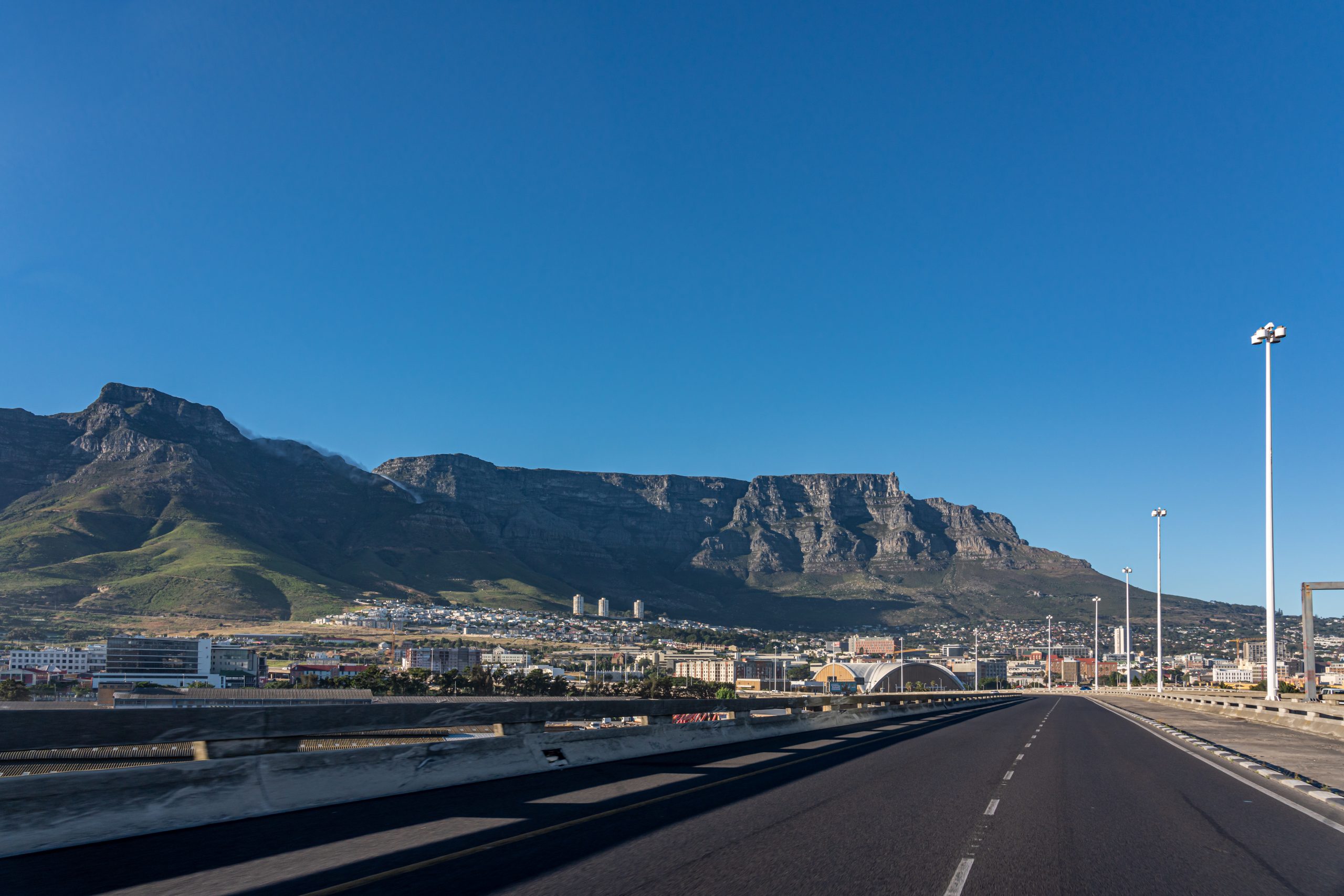 A highway leading toward the city of Cape Town with the massive Table Mountain dominating the background under a clear blue sky.