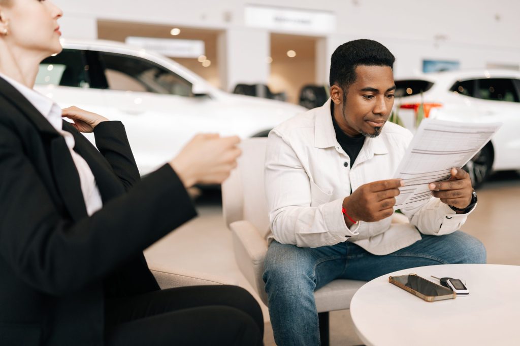A man sitting in a car dealership reviews paperwork at a small table, with a woman beside him adjusting her jacket and several cars visible in the background.