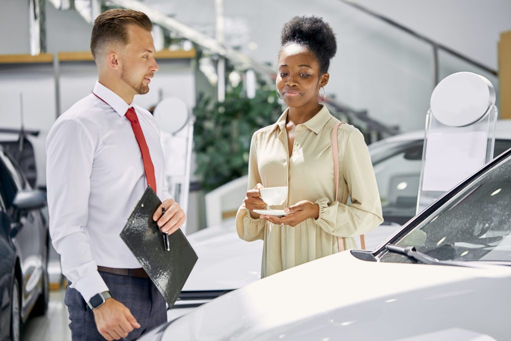 A woman holding a cup talks with a car salesperson who is holding a clipboard as they stand beside a vehicle in a dealership.