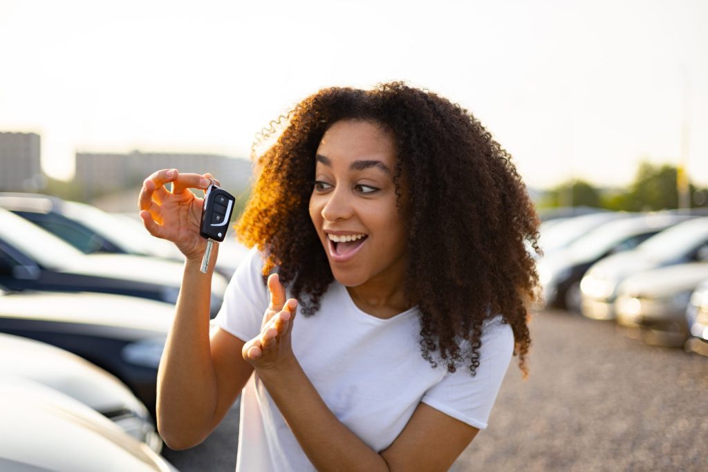 A woman smiling excitedly while holding up car keys in a parking lot full of vehicles.