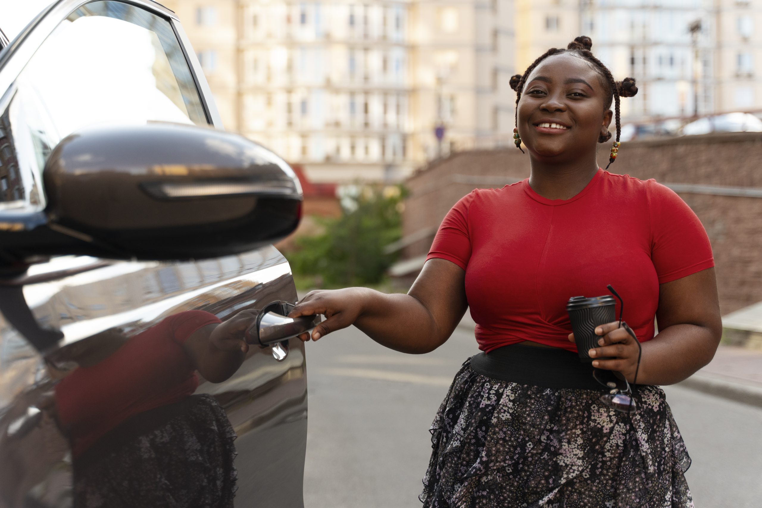 A smiling woman in a red shirt and patterned skirt opening a car door while holding a coffee cup and sunglasses.