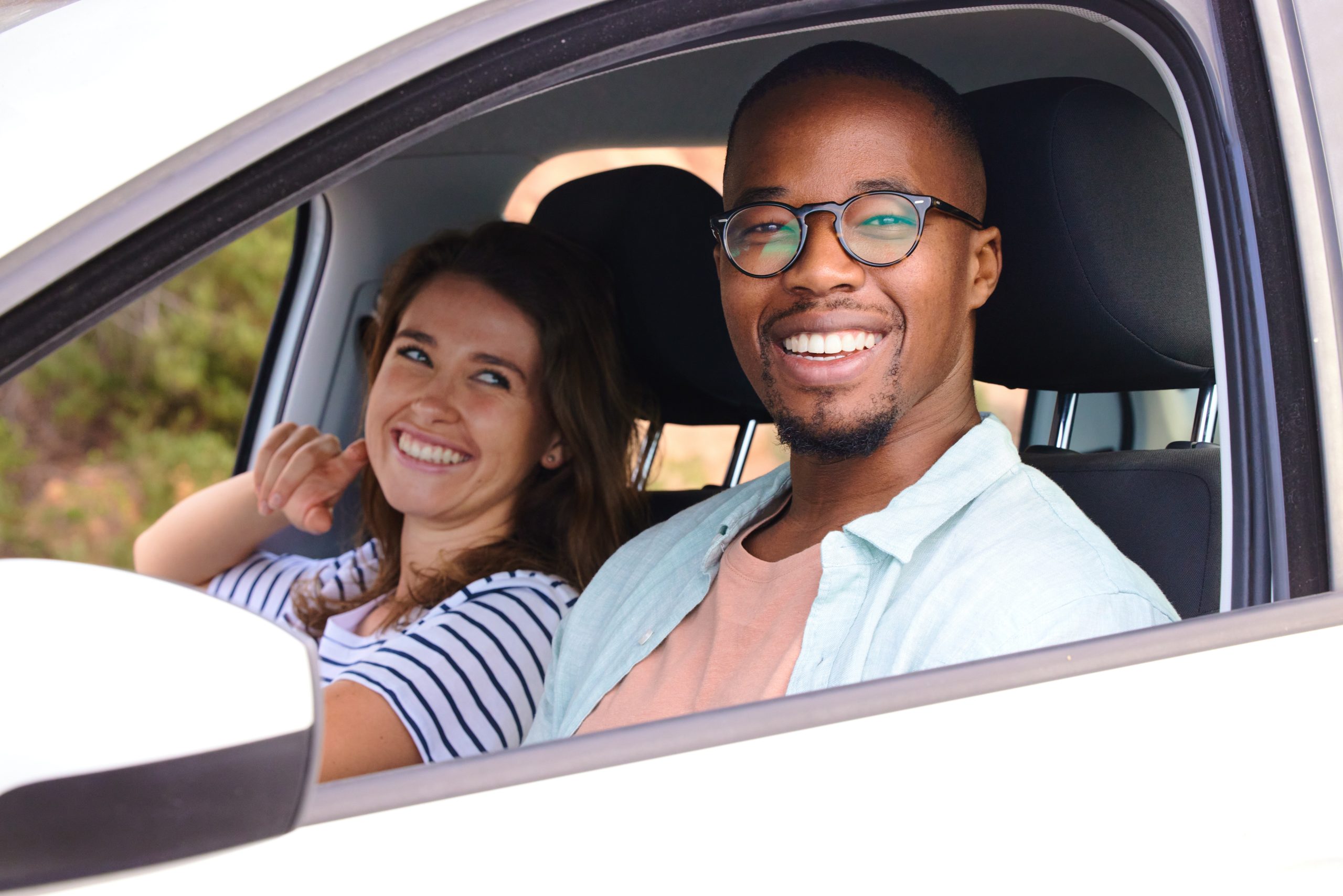 A smiling man in the driver's seat and a woman in the passenger seat of a car, looking out the window.
