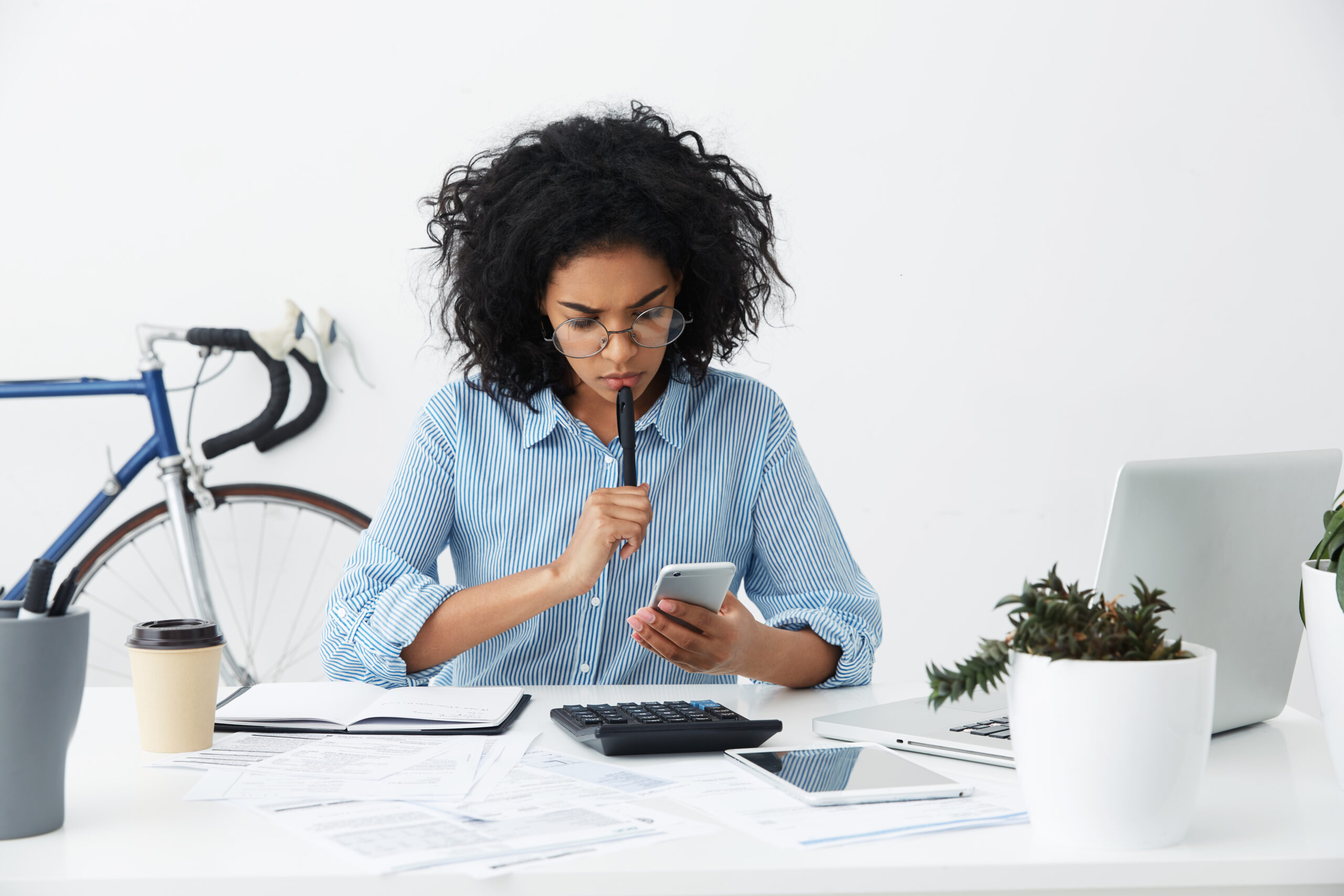 Frustrated young female entrepreneur in formal shirt and eyewear having a problem while working on financial report in office, reading information on mobile phone screen with perplexed expression