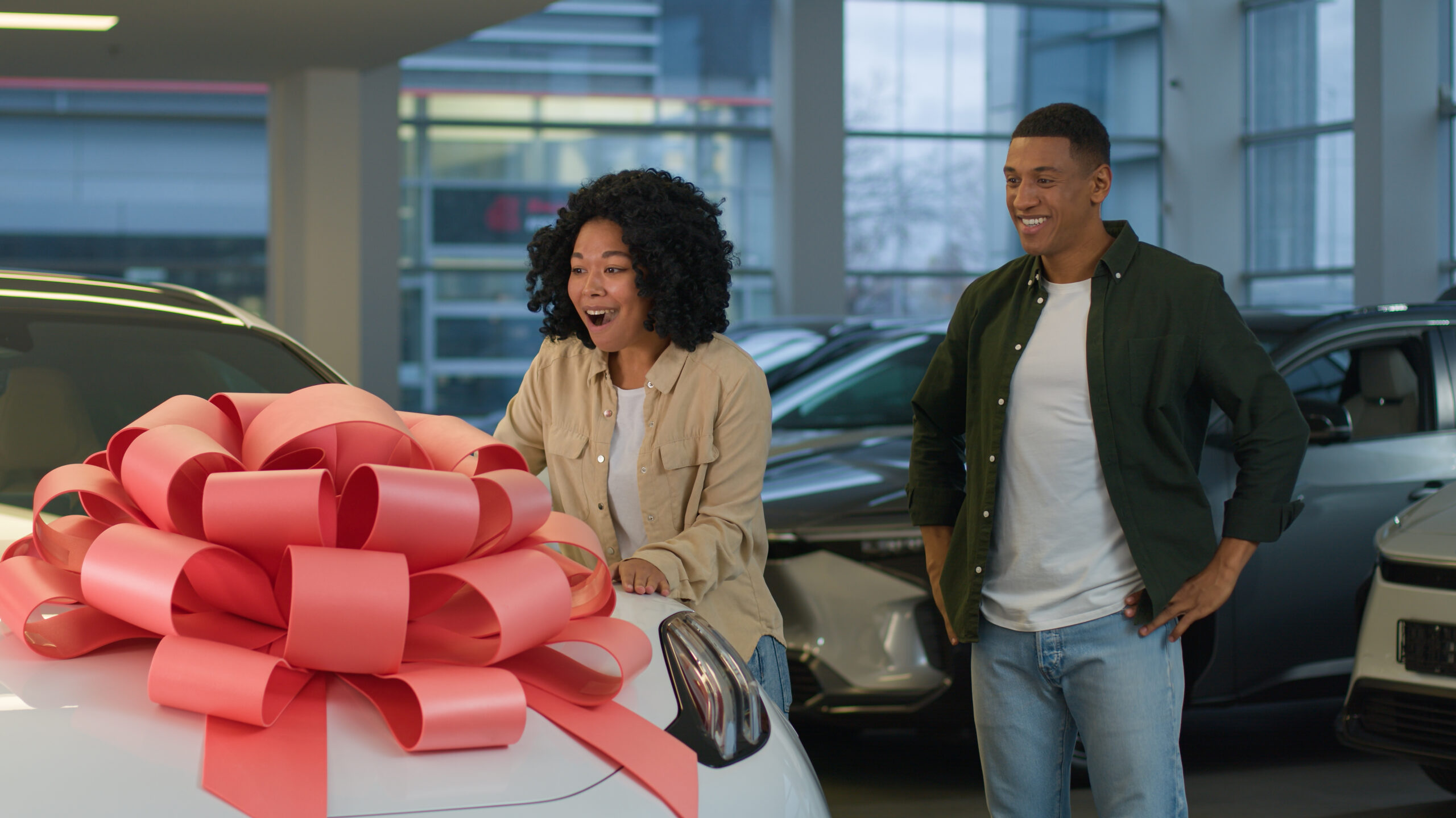 African American couple boyfriend girlfriend in dealership showroom surprise new crossover car gift birthday present with red bow on auto happy woman wife rejoice enjoy luxury automobile hug husband. High quality 4k footage
