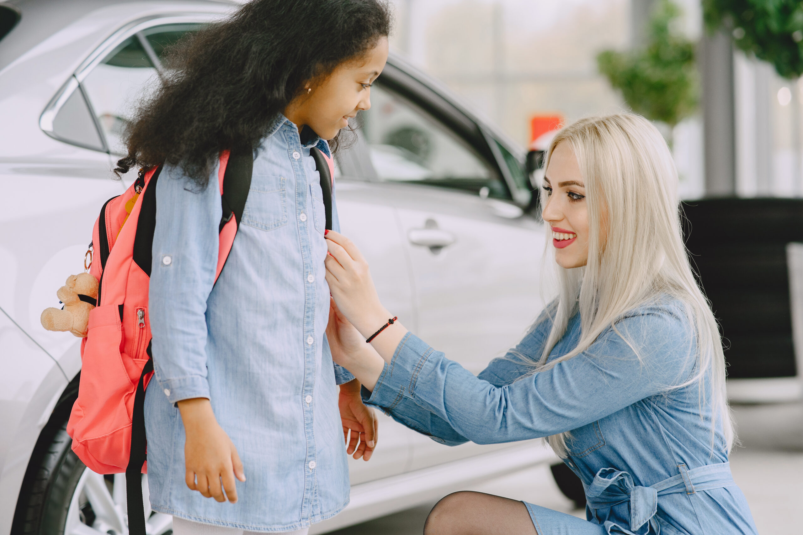 Gemini said A blonde woman in a denim dress smiles while adjusting the pink backpack of a young girl in a denim shirt, with a white car in the background.
