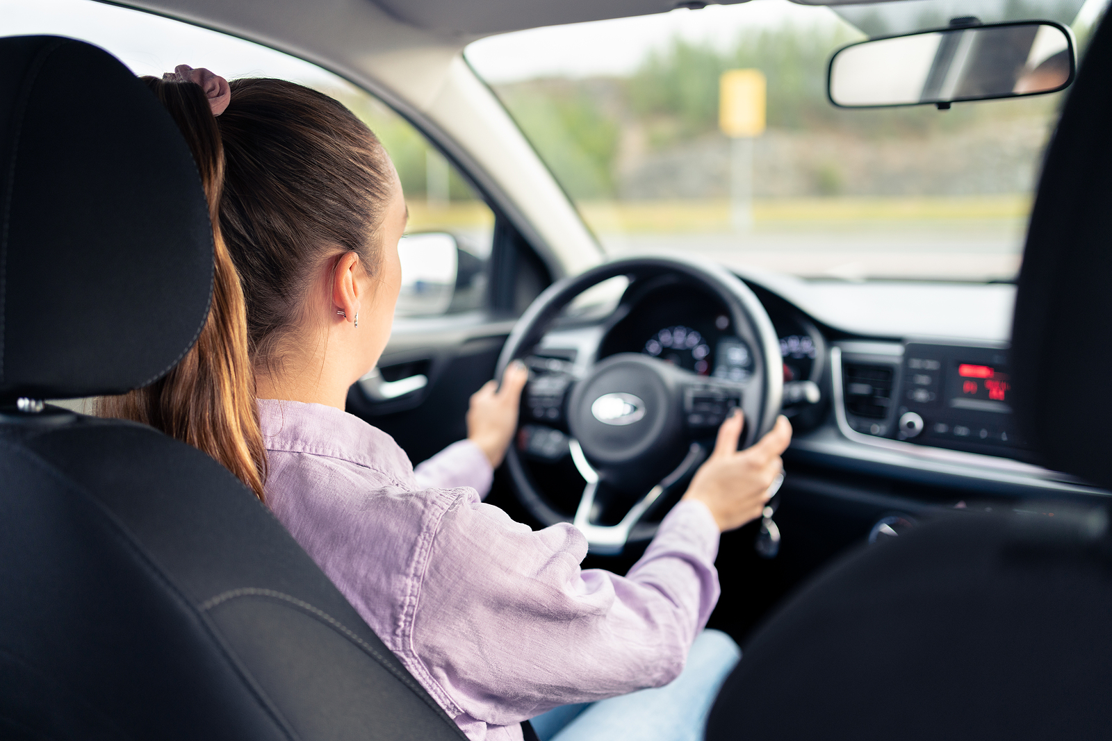 Woman driving car. Young driver. Testing vehicle in dealership or student training in school to get license. Hands on steering wheel. Behind back inside view of cockpit. Rent or ride share service.
