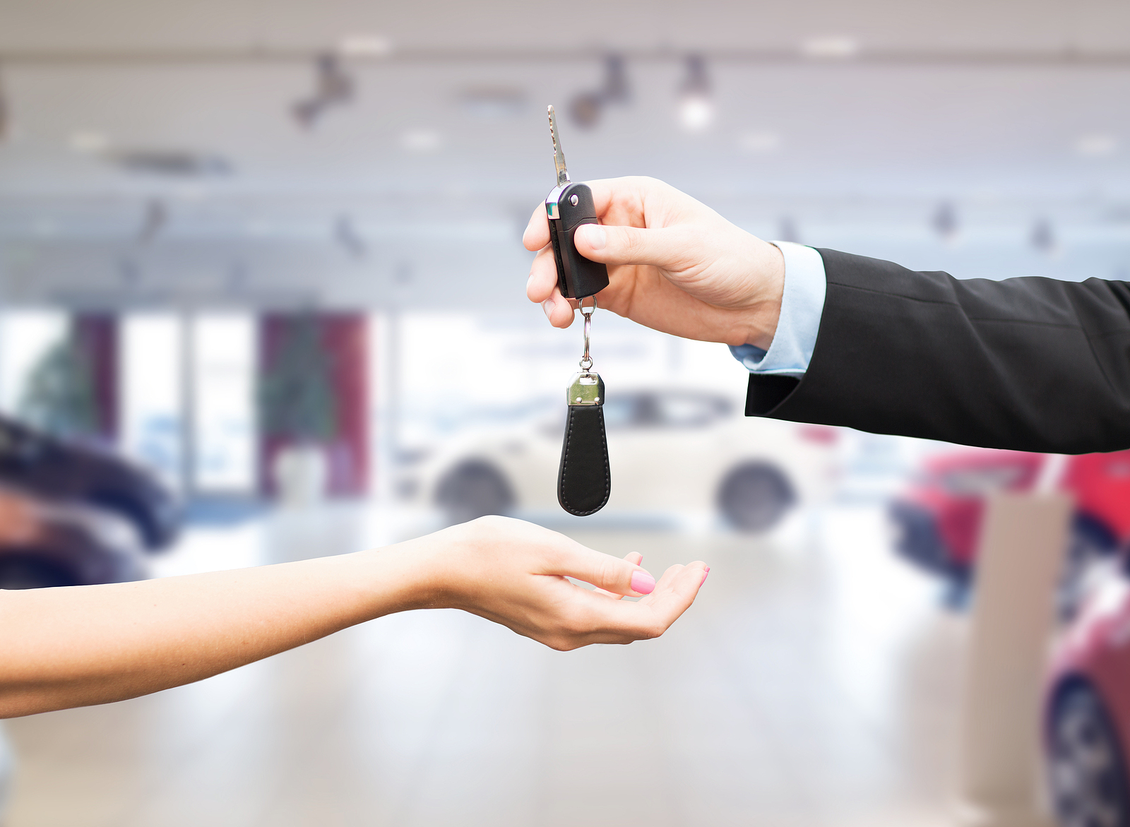 Close-up of a sales representative handing car keys to a customer in a modern, blurred automotive showroom setting.
